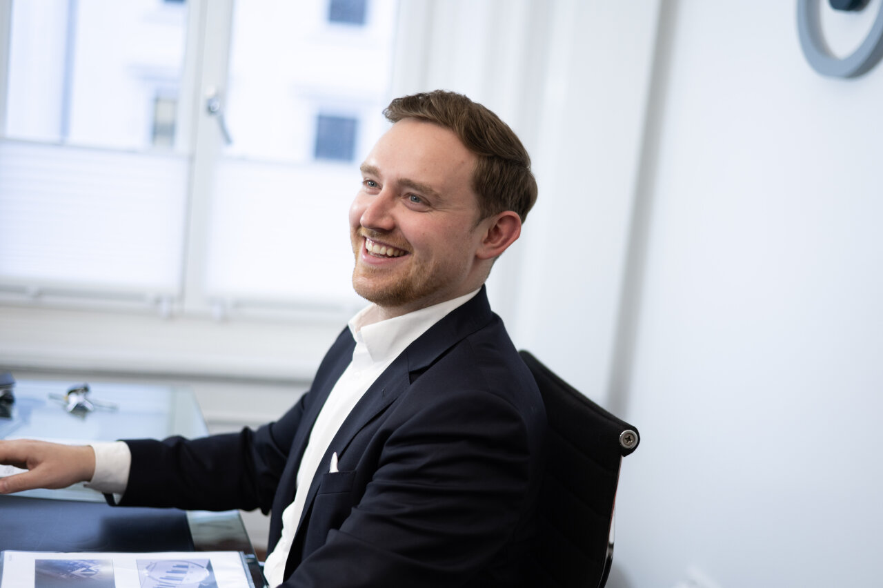 Ein lächelnder EGIDO-Mitarbeiter sitzt an einem Schreibtisch in einem modernen Büro. Er trägt einen dunkelblauen Anzug und hat die Haare nach hinten frisiert, während er auf ein Dokument schaut, das vor ihm liegt.