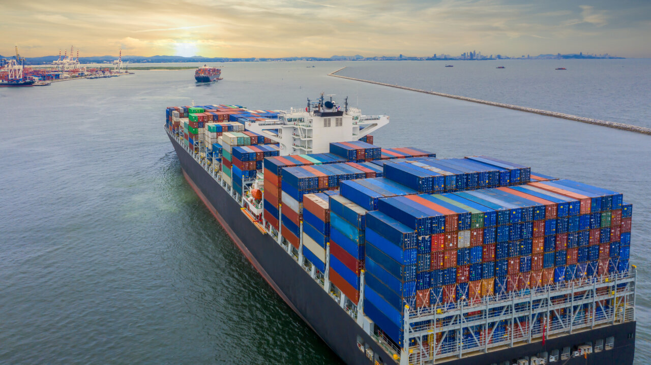 Ein großes Containerschiff fährt durch ruhiges Wasser, beladen mit bunten Containerstapeln. Im Hintergrund stehen Kräne und weitere Schiffe im Hafen, die typische Abläufe der Logistik-Branche zeigen, während die Sonne am Horizont aufgeht.